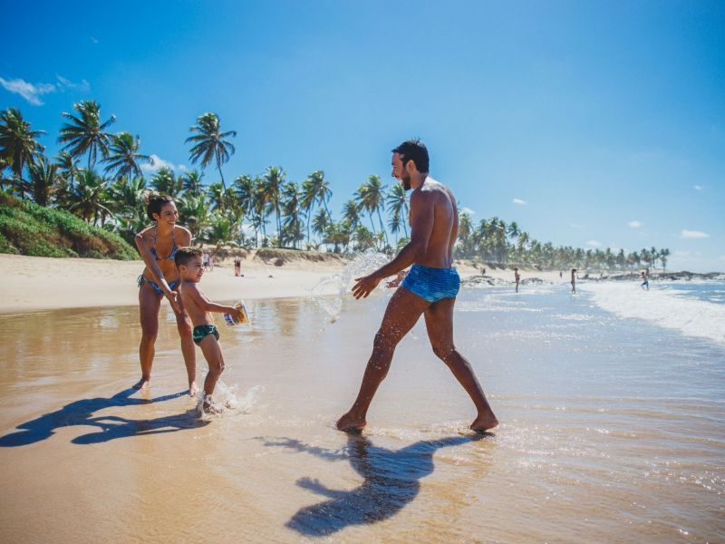 Homem, mulher e criança brincando em uma praia com céu azul próxima a uma pequena vegetação com palmeiras.