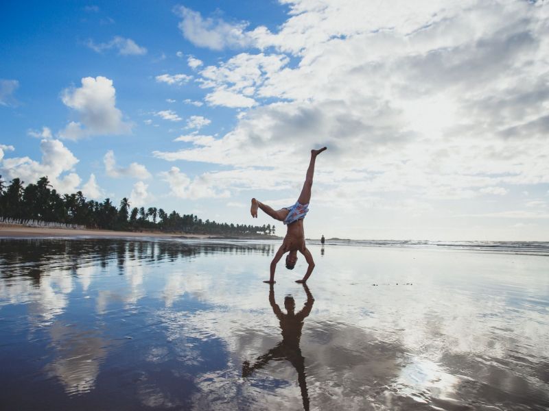 Homem plantando bananeira à beira-mar, em frente a um céu azul com nuvens brancas e próximo a um conjunto de árvores.