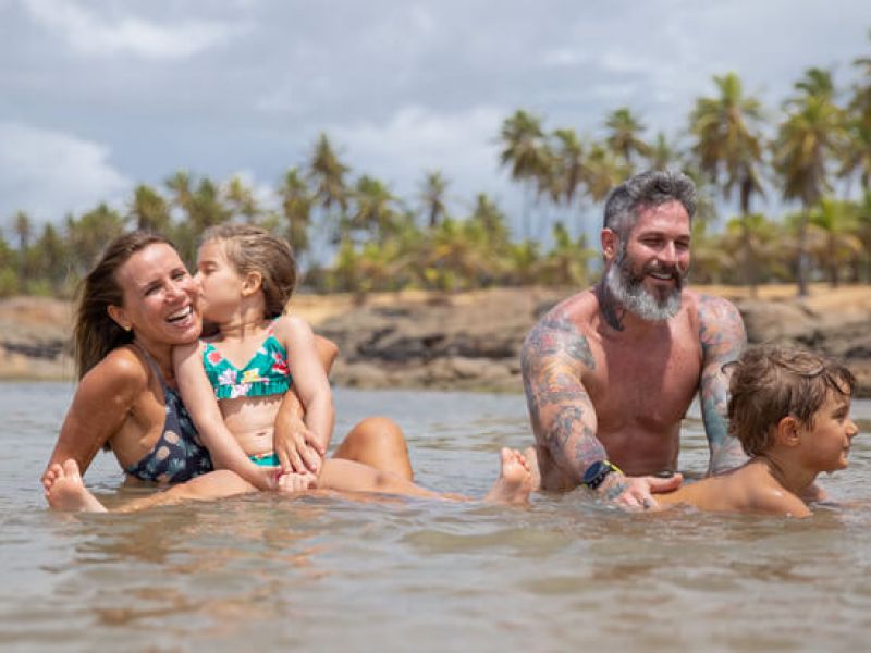 Dois adultos e duas crianças brincando na água de piscinas naturais na Bahia com coqueiros e um céu azul no fundo.
