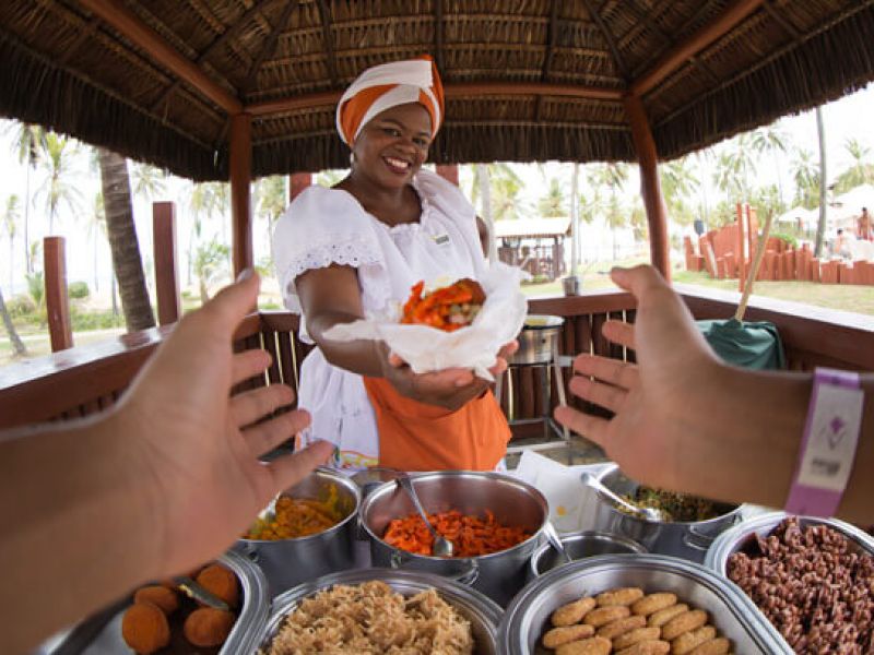 Imagem de uma mulher com uma roupa laranja e branca oferecendo um prato com comidas típicas da Bahia para mãos estendidas.