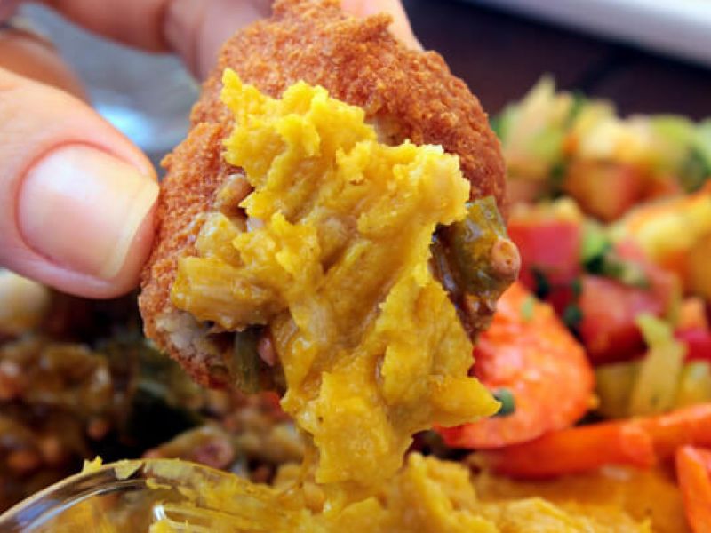 Imagem da receita de acarajé pronta sendo segurada por uma pessoa em frente a um prato cheio de alimentos coloridos
