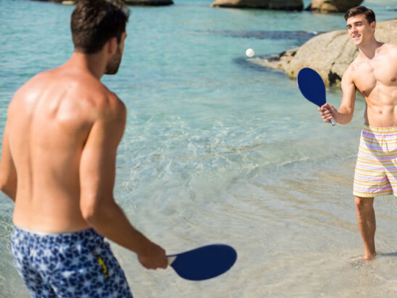 Imagem de dois homens próximos ao mar segurando raquetes e aprendendo a como jogar frescobol na praia.