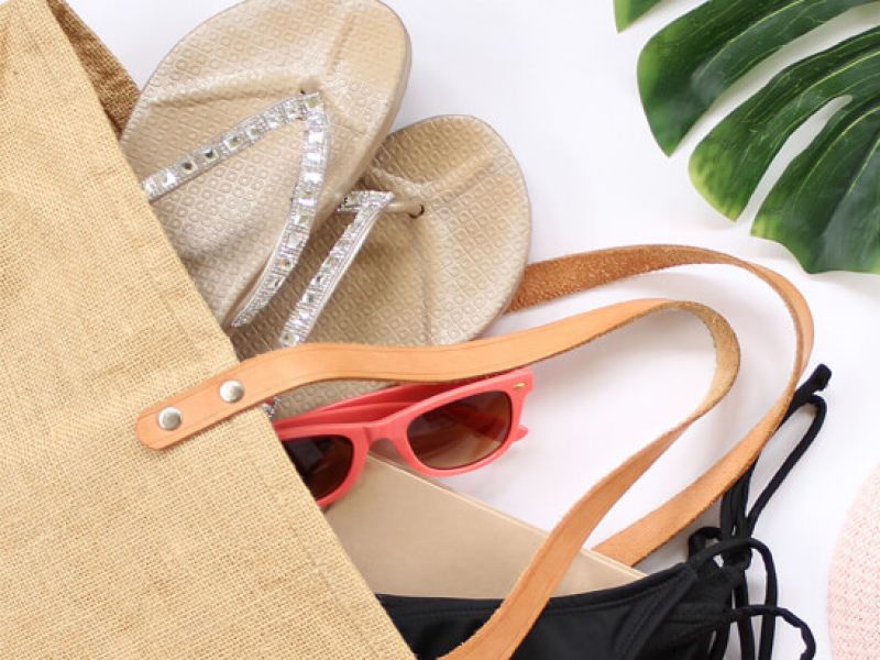 Imagem de bolsa de praia com chinelos, óculos de sol e biquíni. O acessório está num fundo branco enfeitado com uma folha