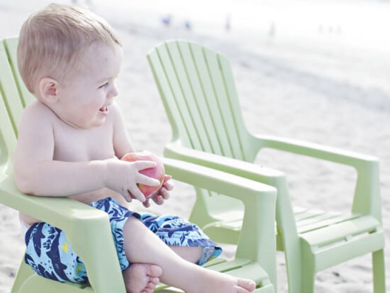 Imagem de bebê sentado em cadeiras verdes numa praia. Ele está sorridente e olhando para o horizonte.