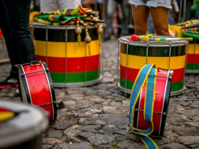 Instrumentos de percussão coloridos no chão enquanto algumas parte das pernas de algumas pessoas estão paradas ao lado dos instrumentos