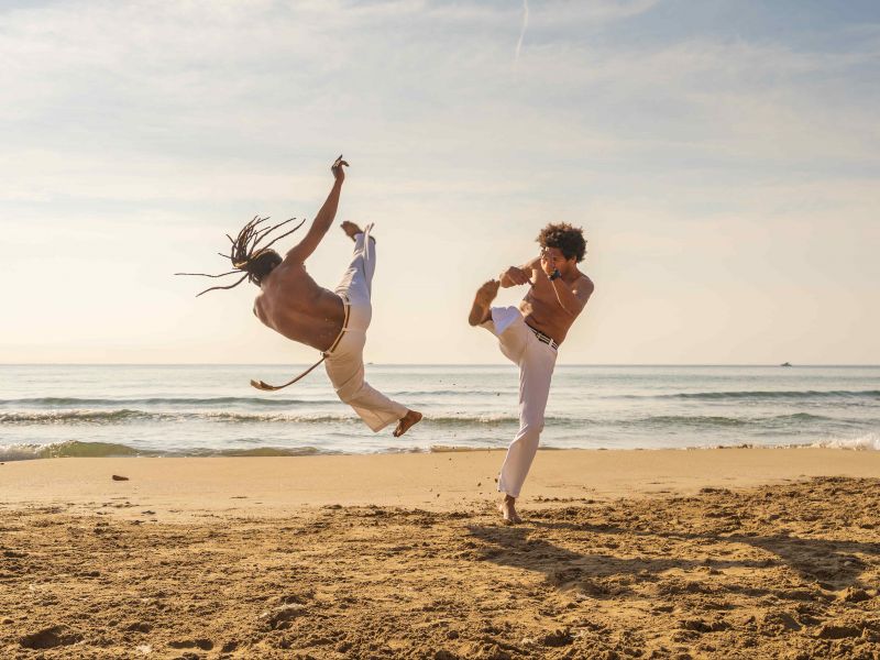 Duas pessoas jogando capoeira na areia da praia com o mar ao fundo