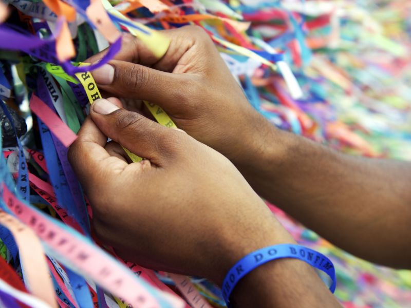 Duas mãos de uma pessoa seguram uma fitinha amarela do Senhor do Bonfim, amarrada no muro da igreja, onde estão milhares de outras fitinhas coloridas