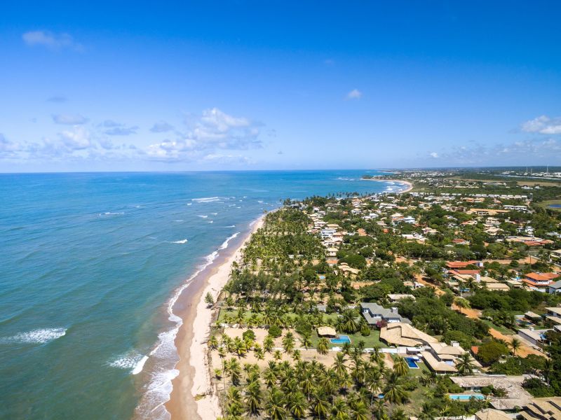 Orla da praia de Itacimirim, com as casas se misturando com a vegetação, enquanto o mar azul em contraste com a faixa de areia