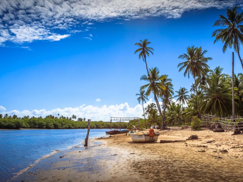 Orla do Sítio do Conde, em Conde Bahia. O mar azul entra em contraste com o a areia branca e os coqueiros verdes. Um barco está parado na areia