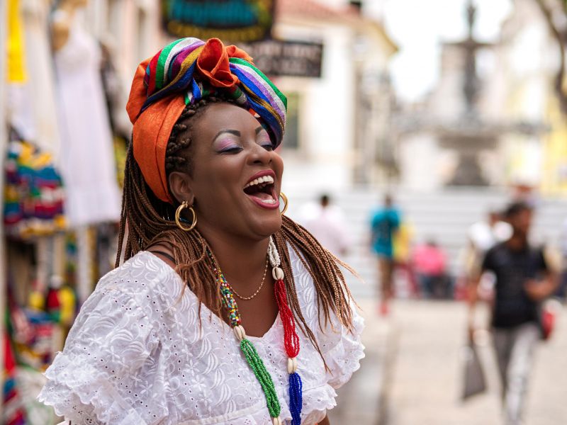 Mulher usando trajes de baiana com um vestido branco, lenço colorido na cabeça e colare de contas