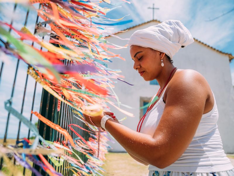 Mulher de branco amarrando fitas do senhor do bonfim em uma grande em frente a uma igreja