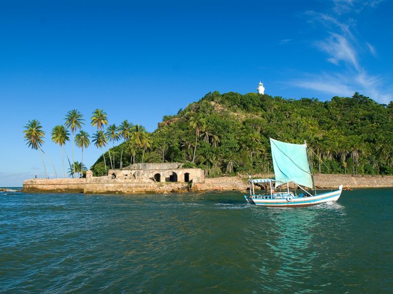 Barco azul navegando no mar com o forte e vegetação de Barra Grande ao fundo