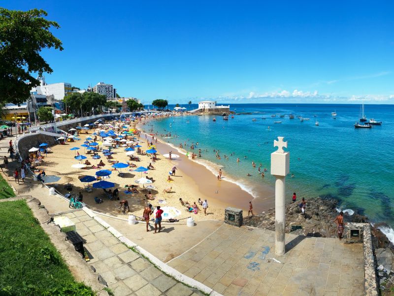 Praia de Porto da Barra e cruz de fundação da região. A areia da praia está chega de pessoas com guarda-sóis coloridos 