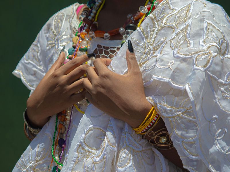 Parte do corpo de uma mulher trajando as tradicionais roupas de baiana, com um vestido branco e muitas contas e pulseiras coloridas