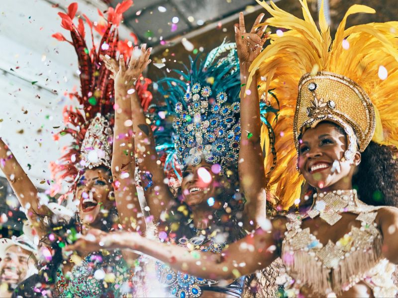 Três mulheres vestidas com os trajes típicos do samba e carnaval carioca, com plumas e brilho