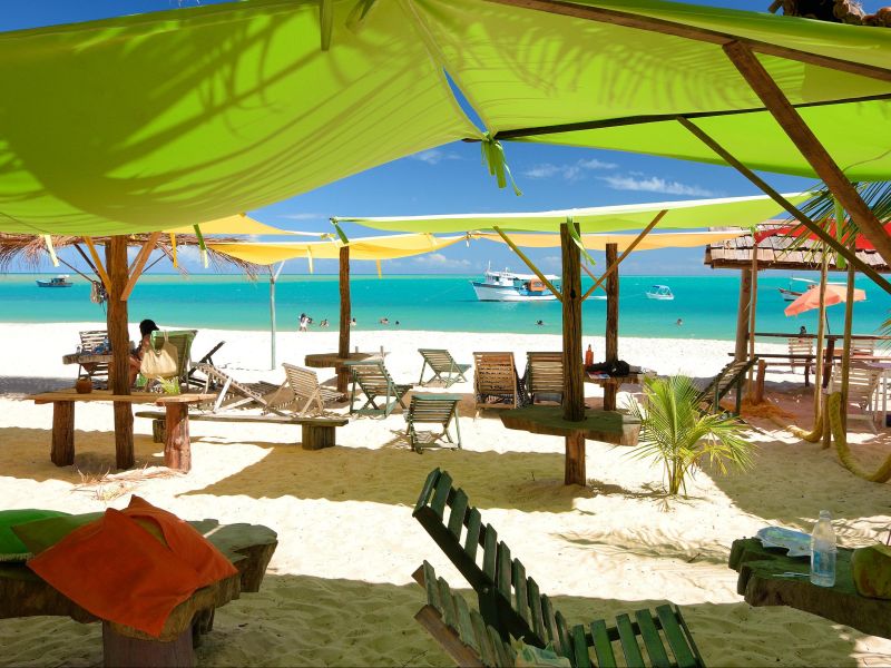 Tendas coloridas sobre bancos de madeira e o mar esverdeado ao fundo, na Praia de Corumbau